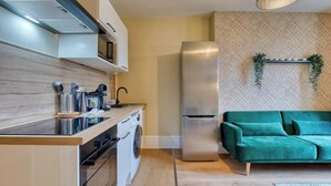 A modern kitchen with a built-in oven, microwave, and stainless steel refrigerator. There is a green sofa and a decorative plant in the living area.