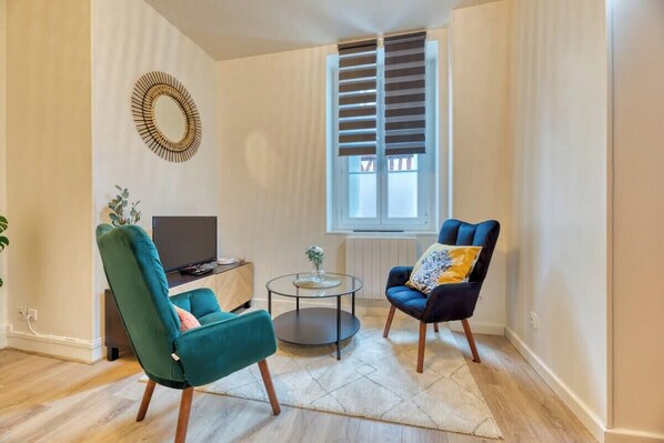 A living room with a teal armchair, a dark armchair with a patterned cushion, a small round coffee table, a TV, and a window with blinds.