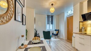 Une cuisine moderne dotée d’un four encastré, un coin repas avec une table dressée pour deux personnes, et un coin salon confortable avec un fauteuil et un canapé.