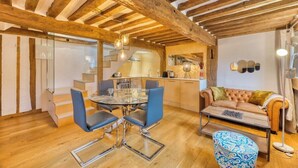 Een moderne keuken met houten balken, een eethoek met een glazen tafel en blauwe stoelen, een bank en een ottoman met een patroon.