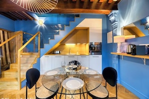 Una cocina moderna con una pared azul de acento, una zona de comedor con mesa y sillas de vidrio, y una escalera con barandillas de madera.