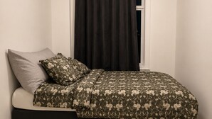 Una cama individual con una colcha estampada, un almohábil gris y un cabecero negro en una habitación con suelo de madera clara y paredes blancas.