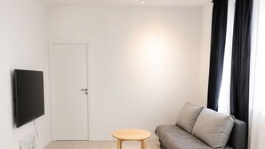 Una sala de estar minimalista con un sofá gris, una mesita redonda pequeña, un televisor de pantalla plana y una puerta blanca.