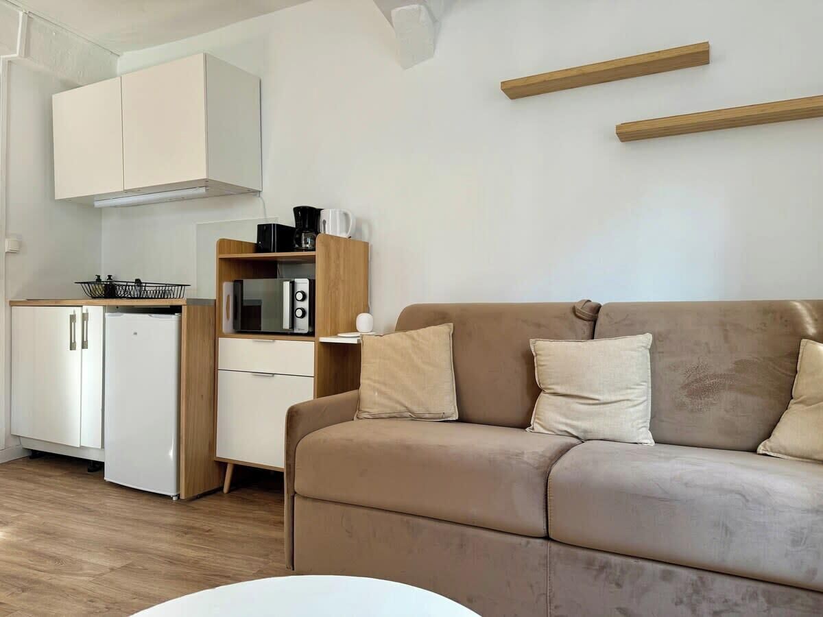 Een moderne keuken met witte kasten, een witte koelkast en een houten aanrecht. Er is een beige bank met kussens in de woonkamer.