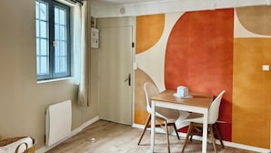 Een eetruimte met een tafel en stoelen, een raam met gordijnen, een radiator en een muur met abstracte oranje en beige patronen.