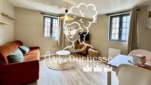 Een gezellige woonkamer met een rode bank, een rond geweven salontafel en een eettafel met stoelen. Er zijn twee ramen met gordijnen, een hanglamp en een klein schapje aan de muur.