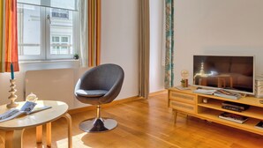 Ein modernes Wohnzimmer mit einem runden Tisch, einem grauen Sessel, einer Holz-TV-Kommode und einem Fenster mit Vorhängen.