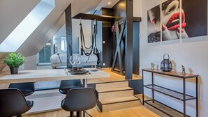 Un espace de salle à manger moderne, aménagé dans un style loft, avec un plafond haut, une table à manger en bois, des chaises noires et une étagère en métal noir agrémentée d’objets décoratifs.