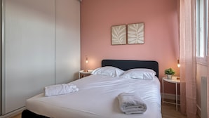 Un lit bien fait, avec du linge de lit blanc et une serviette pliée, une table de chevet avec une lampe et une plante fixée au mur.