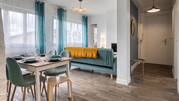 Un salon moderne avec un coin repas, comprenant un canapé, une table dressée pour quatre personnes et une grande fenêtre avec des rideaux.