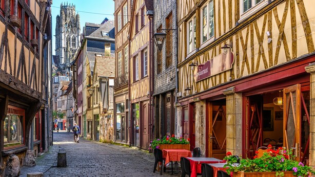 Een geplaveid straatje met historische vakwerkhuizen, een restaurant met terras, en een kerk op de achtergrond.