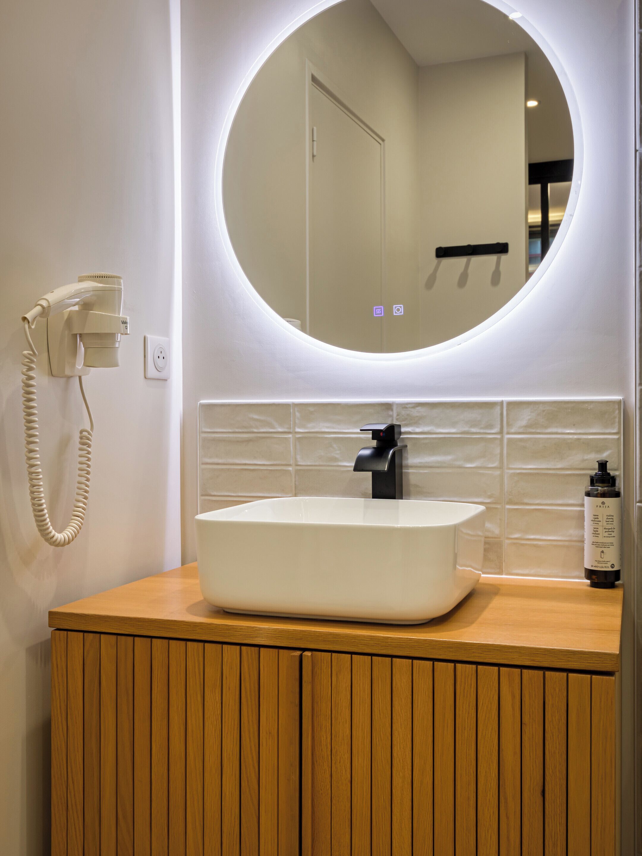 A bathroom with a white sink, a round mirror with a light, and a hairdryer mounted on the wall.
