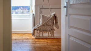 Une pièce avec un plancher en bois, un fauteuil suspendu, un radiateur et une fenêtre donnant sur des immeubles.