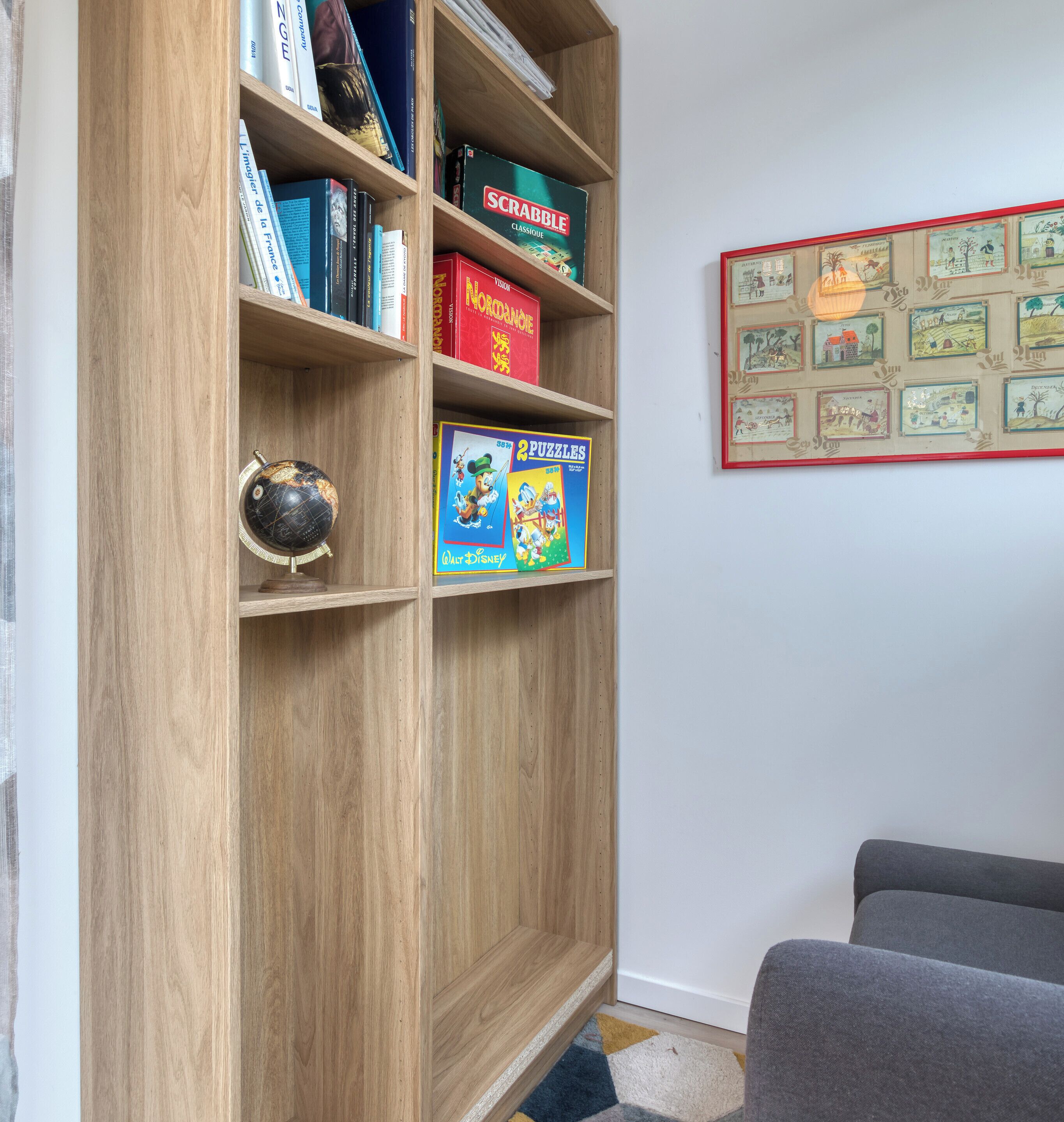A wooden bookshelf with various books and games, a framed picture on the wall, and a grey couch.