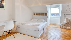 Une chambre à coucher comprenant un lit, un bureau, une chaise, un fauteuil suspendu et une fenêtre donnant sur un paysage agréable.