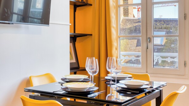 Un comedor con una mesa dispuesta para dos personas, sillas amarillas, un televisor montado en la pared y una ventana con vistas a un edificio y una grúa.