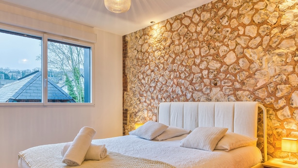 Une chambre avec un mur en pierre, une grande fenêtre et un lit doté d’une tête de lit et de coussins blancs.