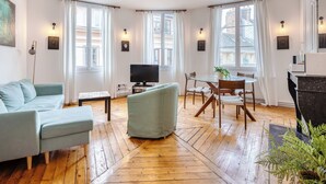 Una sala de estar moderna con un sofá, un televisor, una mesa de comedor con sillas y ventanas amplias con cortinas.