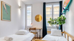 Habitación de hotel con dos camas individuales, un escritorio, una silla, un ventanal grande con cortinas y una planta en maceta.