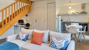A modern apartment with a staircase, a living room with a sofa and cushions, a kitchen with white appliances, and a dining area with a table and chairs. - Le Nicole, un appartement en duplex avec balcon et place de parking. (Rouen)