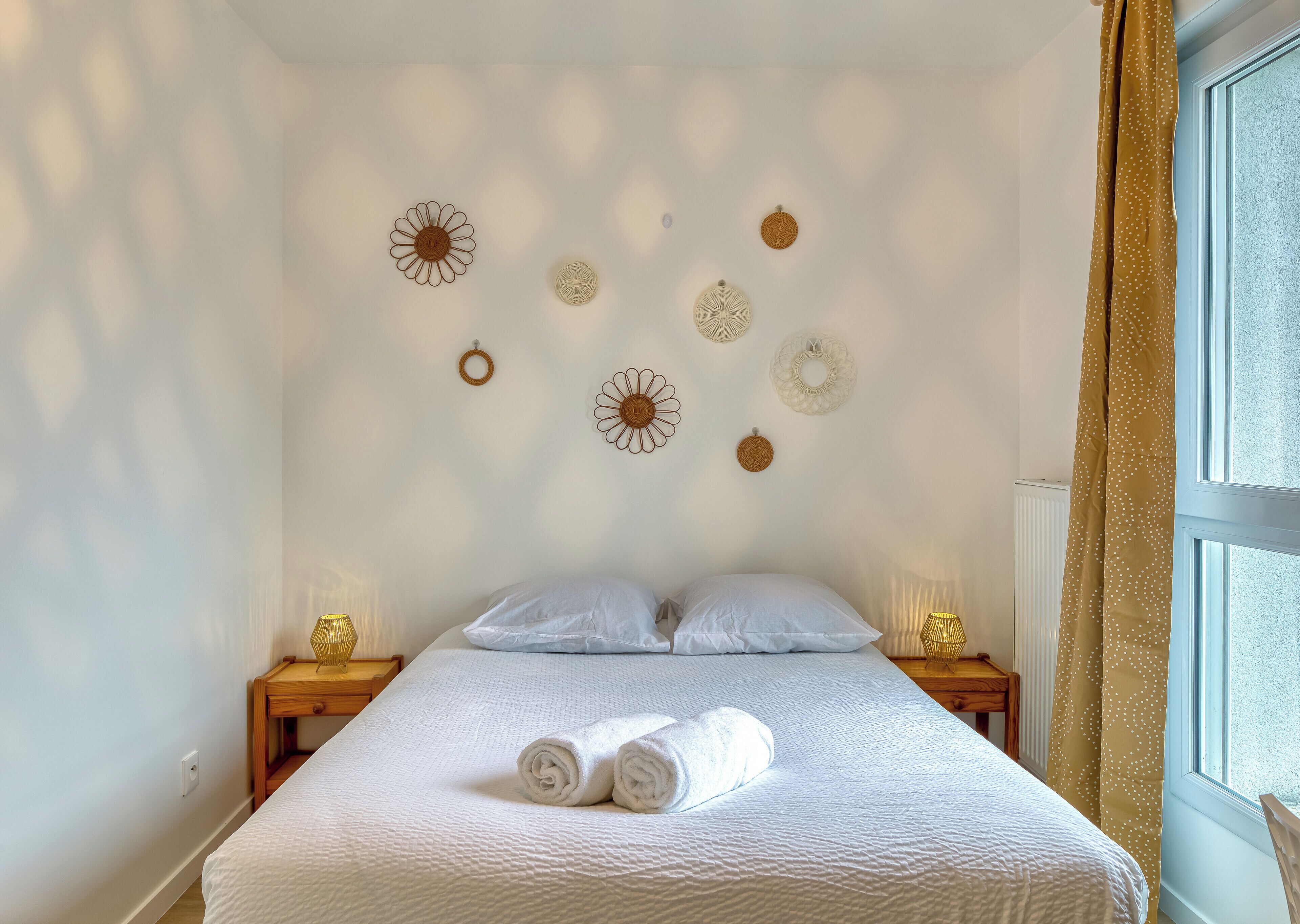 A neatly made bed with white linens and two rolled towels, a wooden nightstand with a lamp, and decorative wall art.
