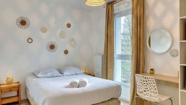 A bedroom with a bed, bedside table, chair, and a window with curtains. - Le Nicole, un appartement en duplex avec balcon et place de parking. (Rouen)