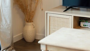 A room with a television on a wooden cabinet, a white table, and a vase with dried plants. - Le Nicole, un appartement en duplex avec balcon et place de parking. (Rouen)