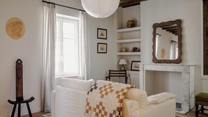 Une pièce chaleureuse avec un canapé blanc, une chaise en bois, une lampe suspendue et une cheminée agrémentée d’un miroir.