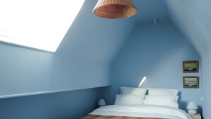 Une chambre avec un plafond en pente, un lit recouvert d’une literie blanche, un plaid rouge à motifs et deux tableaux encadrés accrochés au mur.