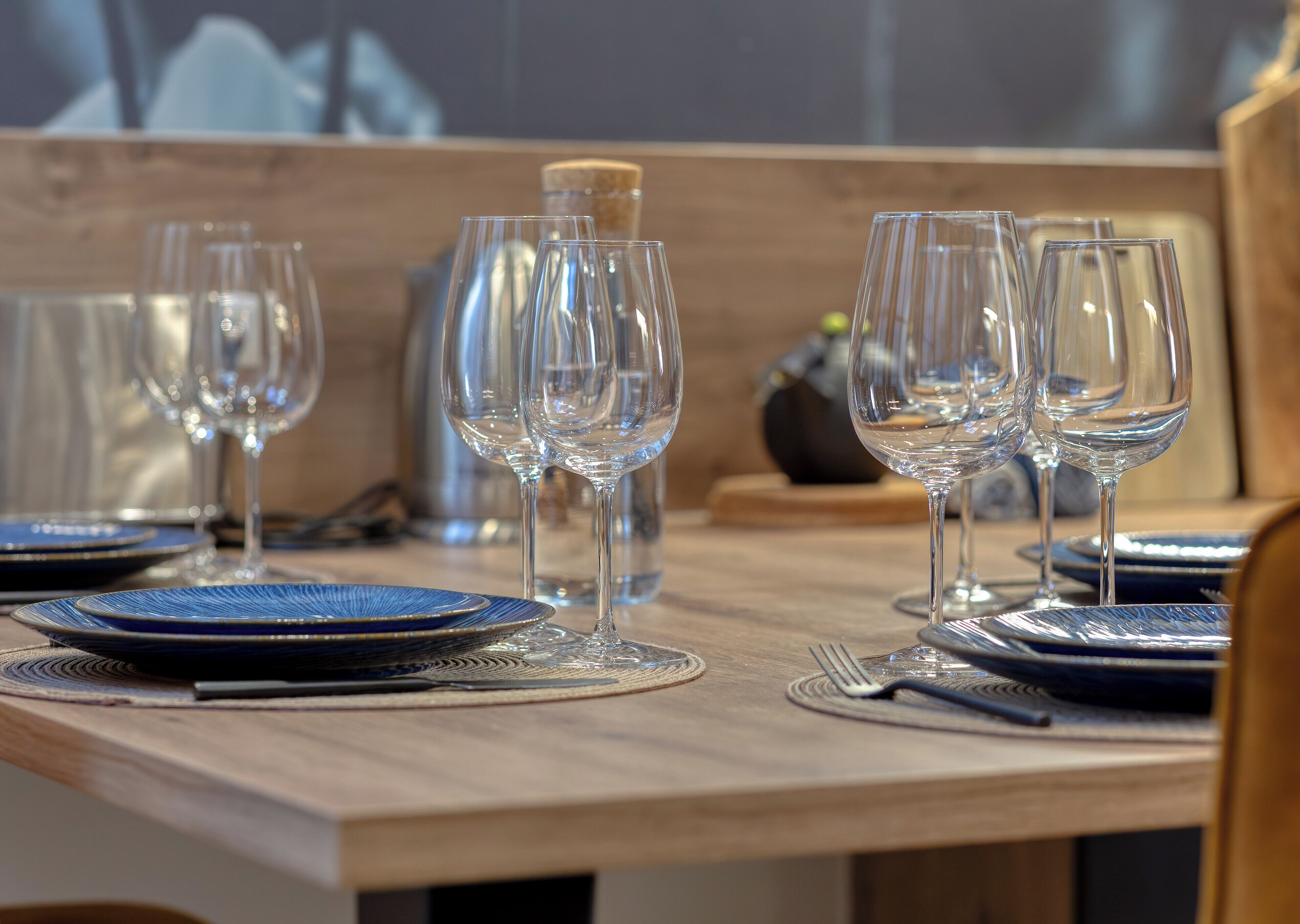 Una mesa de comedor dispuesta con copas de cristal, platos azules y un sacacorchos.