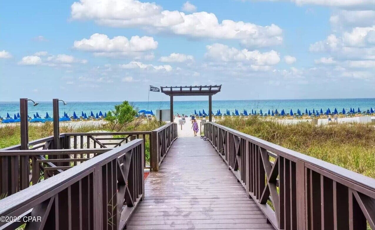 Une promenade en bois mène à une plage où se trouvent des parasols et un panneau indiquant la zone réservée aux baigneurs.