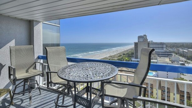 A balcony with a table and chairs, overlooking a beach and cityscape.