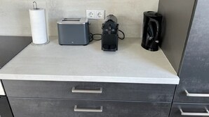Una kitchenette con un tostador, una cafetera y una tetera sobre una encimera. Hay dos enchufes eléctricos y un dispensador de toallas de papel.