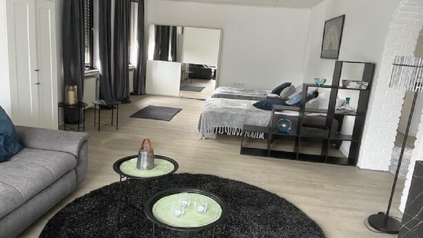 Una habitación de hotel moderna con un sofá gris, un tapete redondo negro, una cama con cabecera gris, una mesita de noche con lámpara y un espejo grande en la pared.