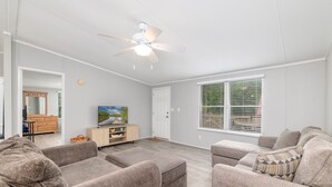 A spacious living room with a ceiling fan, a large window with blinds, and a television on a wooden cabinet.
