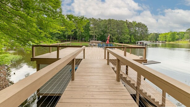 A wooden deck leading to a dock over a calm lake, surrounded by lush greenery and a clear sky.