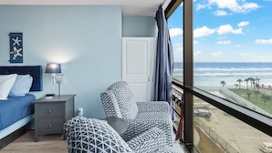 A bedroom with a bed, nightstand, chair, and a view of the ocean. - Oceanside Inn Studios by Brightwild (Daytona Beach Shores)