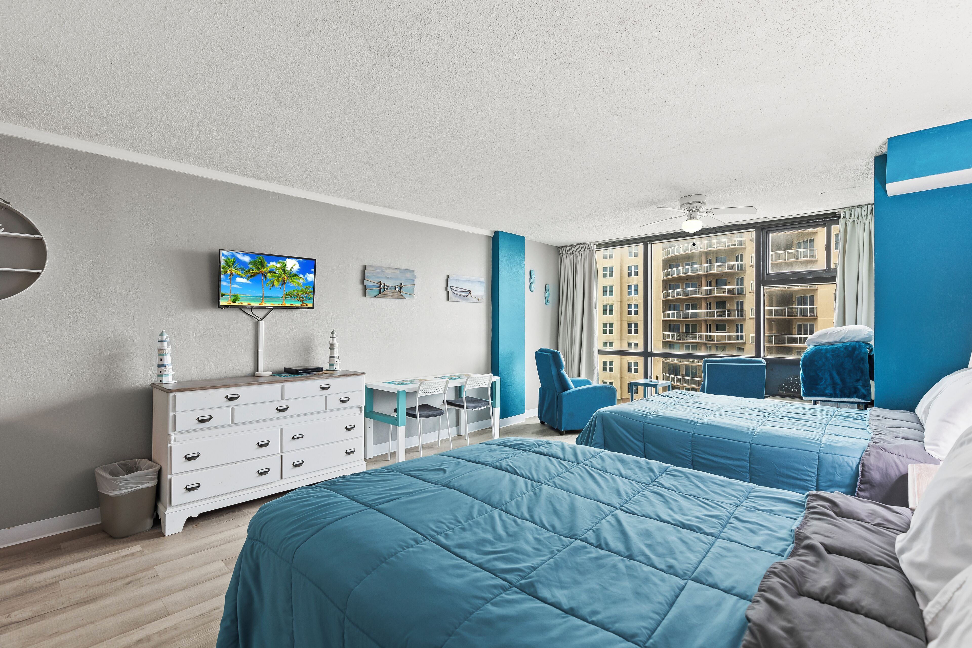 Ein Hotelzimmer mit zwei Betten, einem Schreibtisch mit Stühlen, einem Fernseher und Blick auf ein Gebäude durch große Fenster.