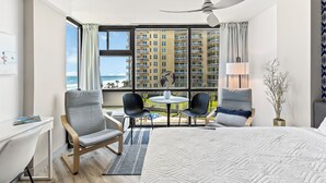 A hotel room with a bed, a desk, two chairs, a table, a ceiling fan, and a large window with a view of a high-rise building and the ocean. - Oceanside Inn Studios by Brightwild (Daytona Beach Shores)