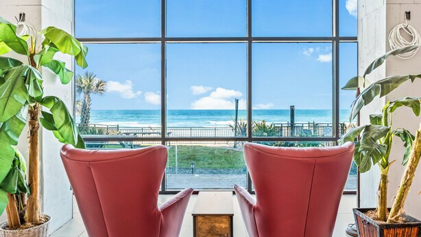 A room with large windows offering a view of the ocean and a bridge. There are two red chairs and potted plants. - Oceanside Inn Studios by Brightwild (Daytona Beach Shores)