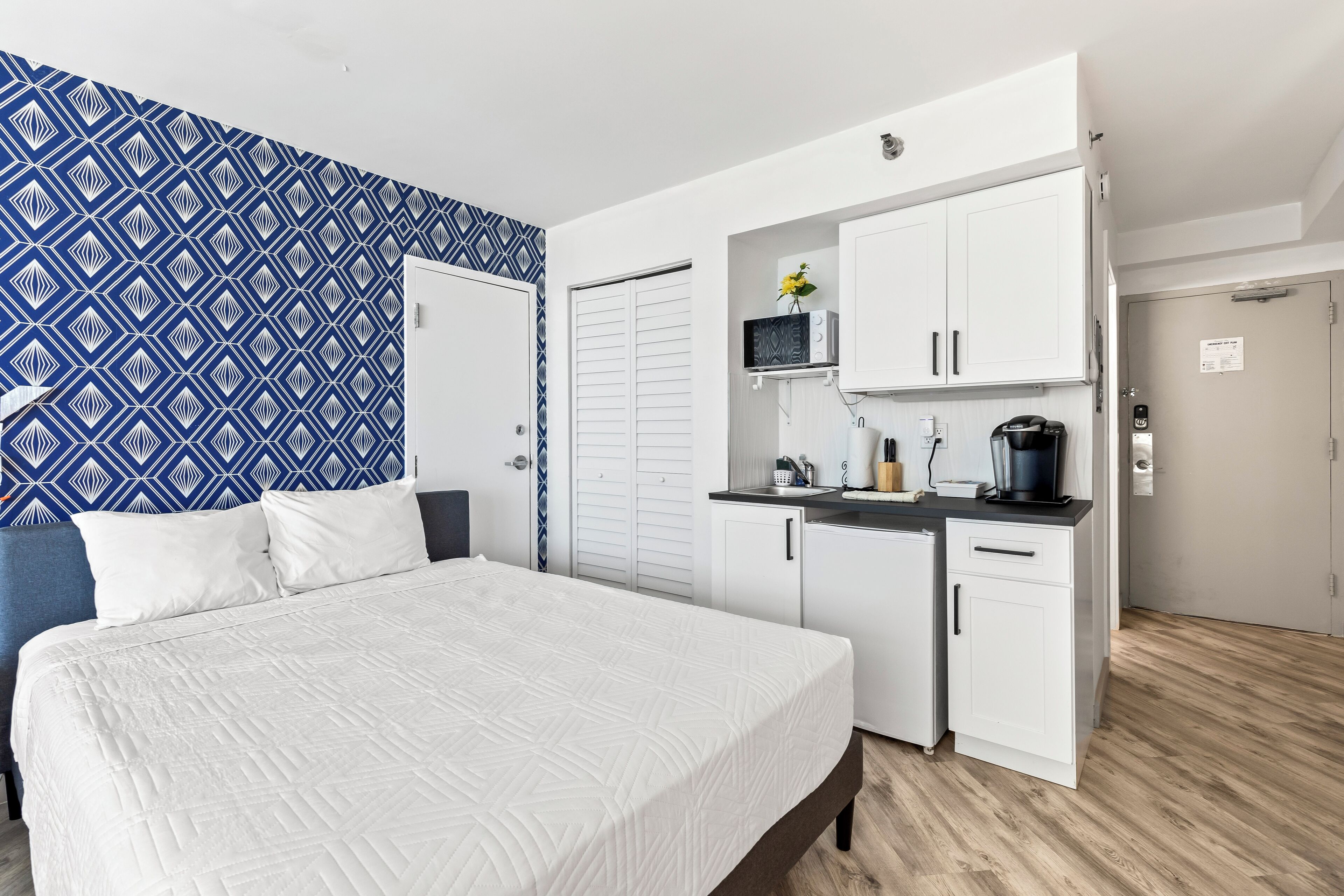 A bedroom with a bed, a wardrobe, a kitchenette, and a patterned wall.