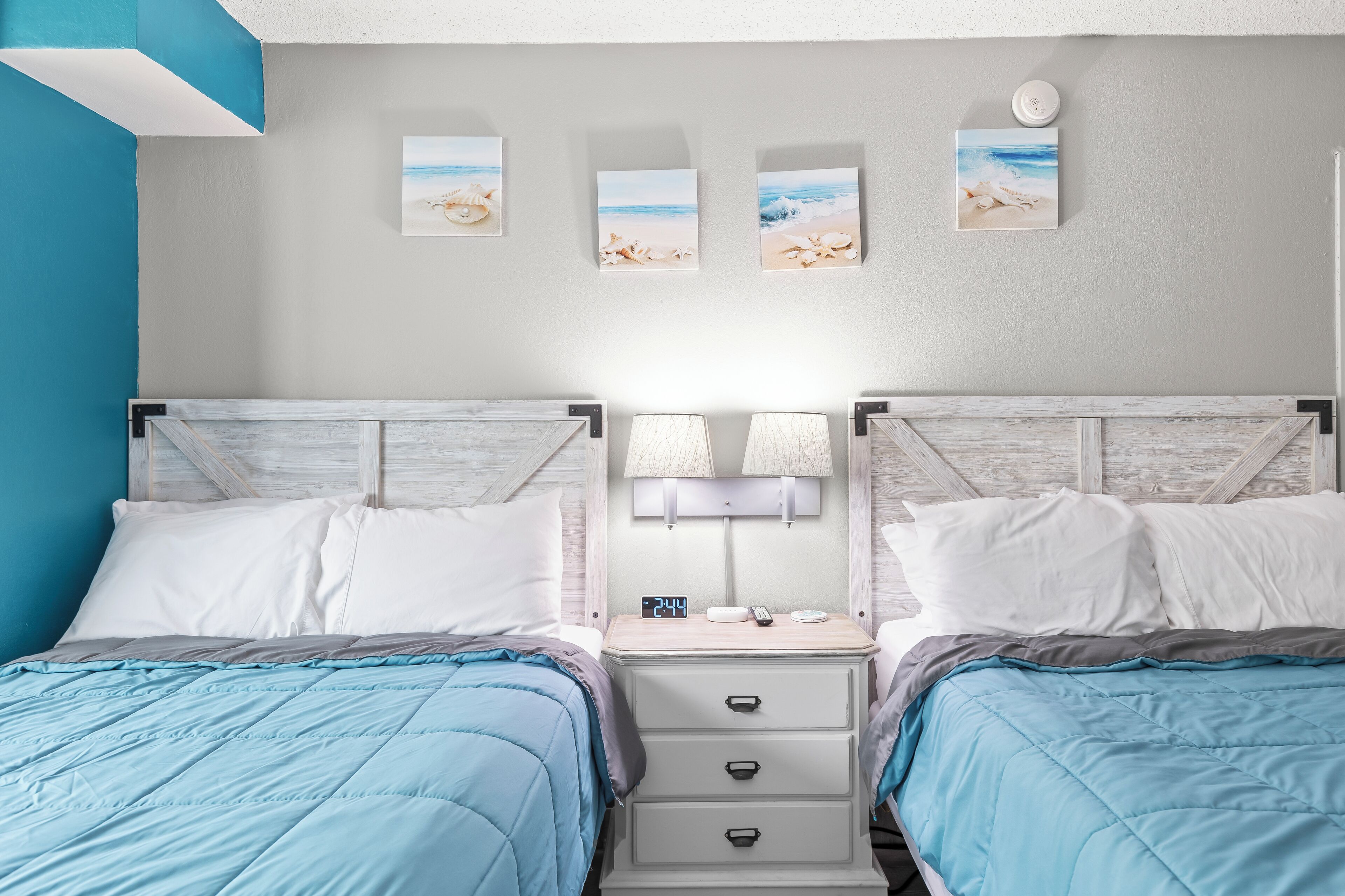 Un dormitorio doble con dos camas, una mesita de noche con lámpara y tres obras de arte con motivos playeros.