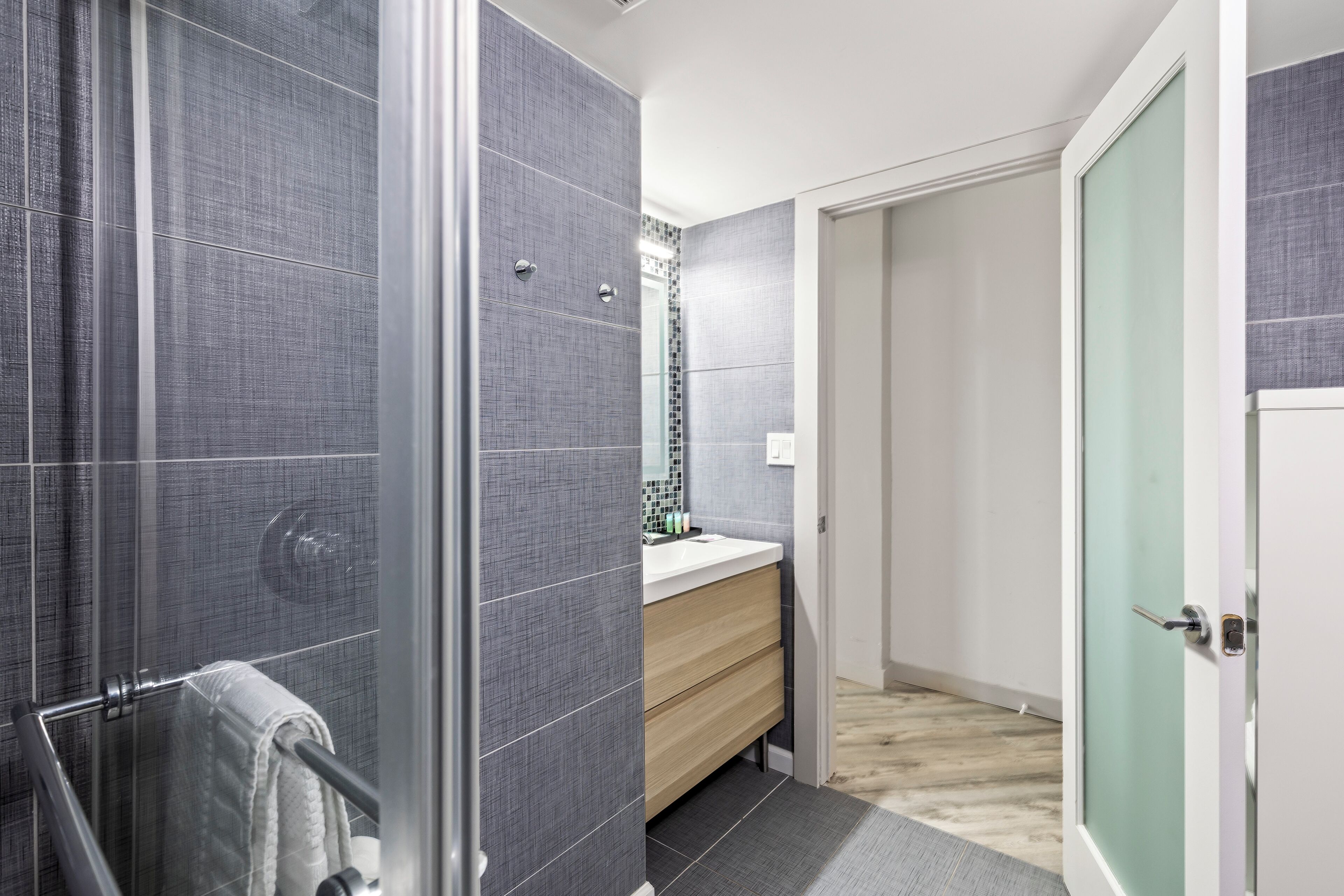 Un bagno moderno con piastrelle grigie, cabina doccia in vetro, mobile bagno in legno e una porta bianca.