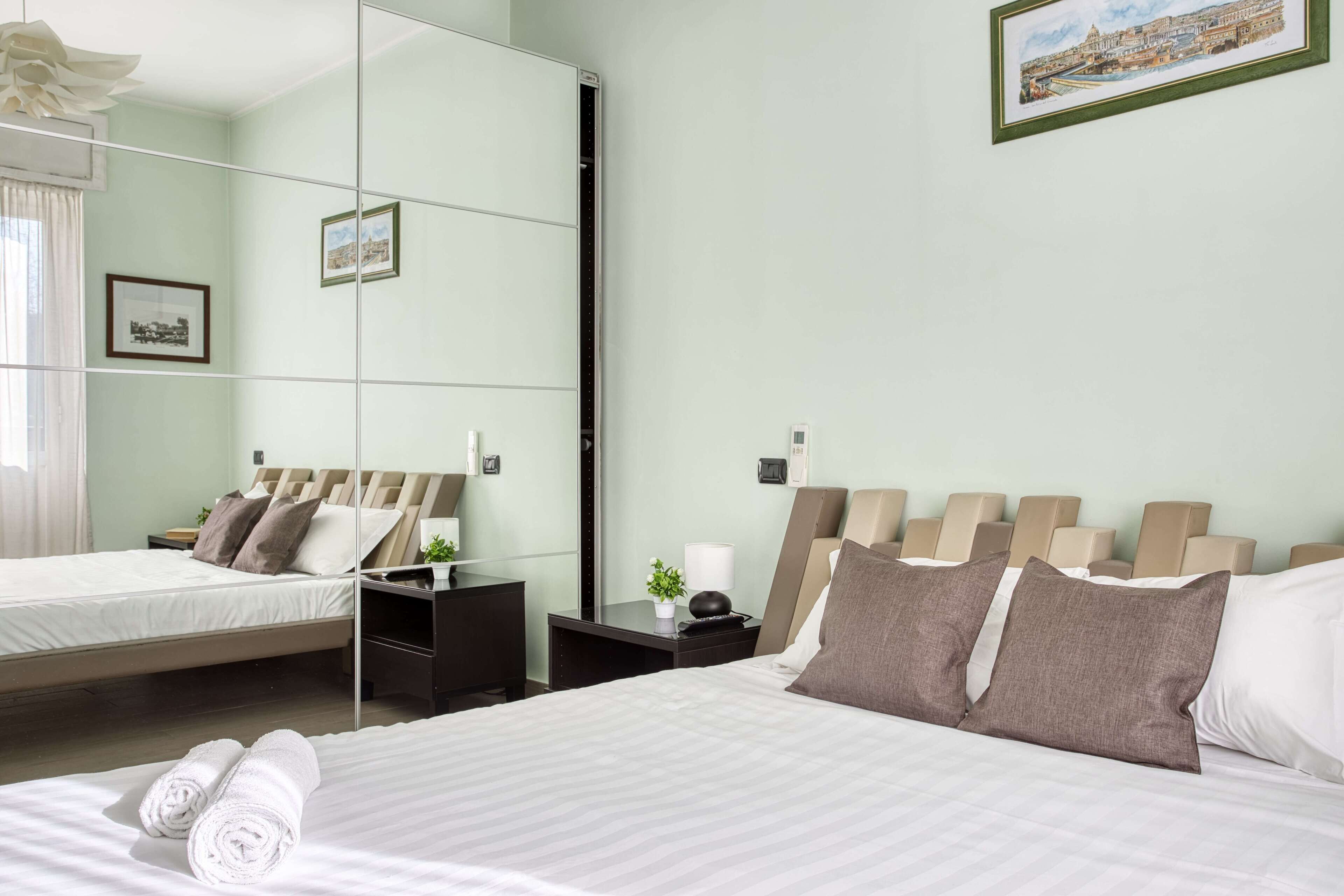 A modern hotel room with a large bed, a bedside table with a lamp, a mirror, and a framed picture on the wall.