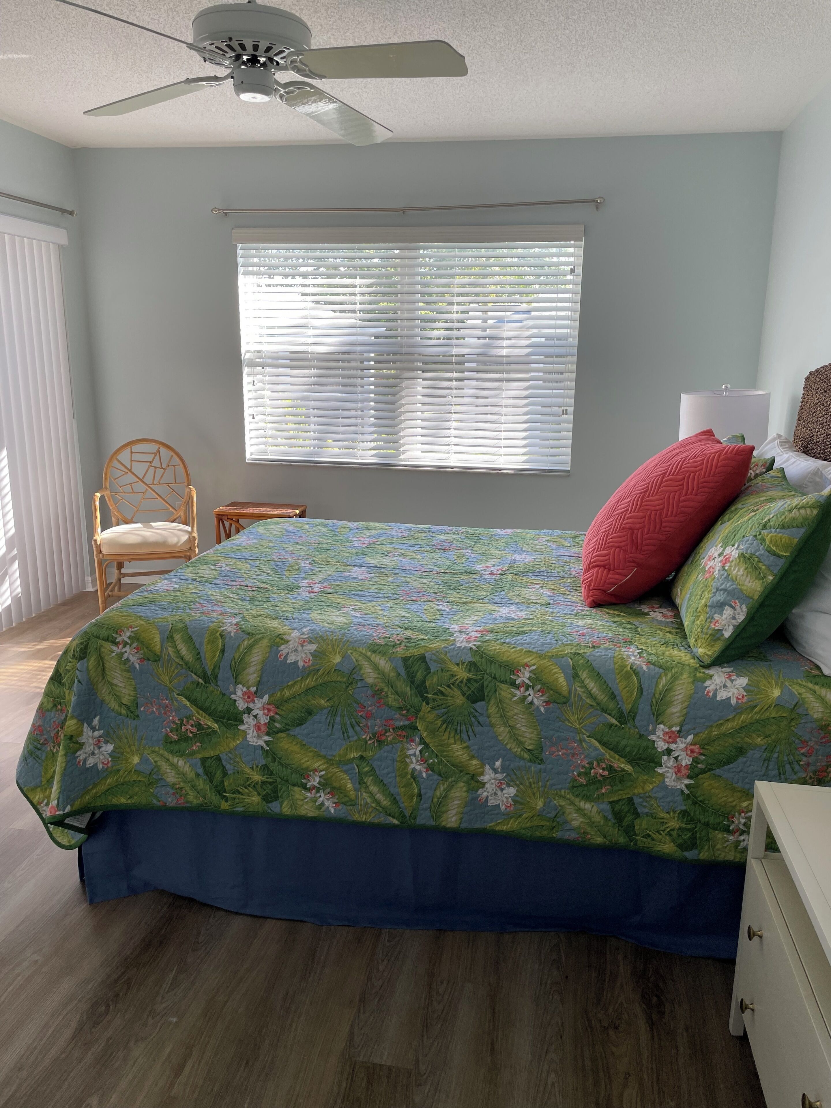 Ein Schlafzimmer mit Bett, Deckenventilator, Fenster mit Jalousien, Stuhl und Nachttisch.