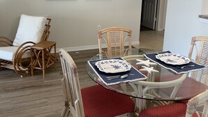 Un coin repas avec une table ronde en verre, des chaises en osier dotées de coussins rouges et deux tableaux représentant des voiliers accrochés au mur.