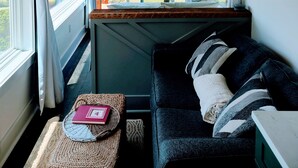 A cozy room with a large window, a dark blue sofa, a woven pouf, and a decorative chandelier.