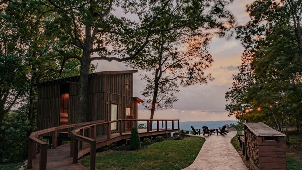 A wooden cabin with a deck and a pathway leading to a seating area with chairs. - The Artistree House A Treehouse in Rising Fawn (Rising Fawn)