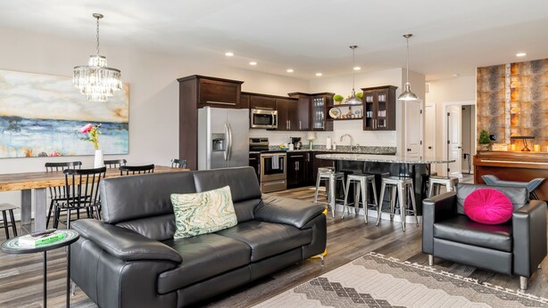 A modern living room with a black leather sofa, a dining table with chairs, a kitchen with dark cabinets, stainless steel appliances, and a bar area with stools. - Highlands Lookout-3BR 3.5BA-Walk to Tons +Parking (Louisville)
