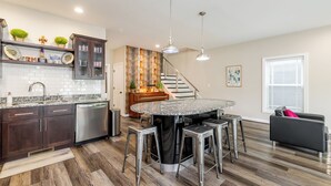 A modern kitchen with dark wood cabinets, a granite countertop, and stainless steel appliances. There is a round dining table with bar stools and pendant lighting. A staircase leads to a second floor. - Highlands Lookout-3BR 3.5BA-Walk to Tons +Parking (Louisville)